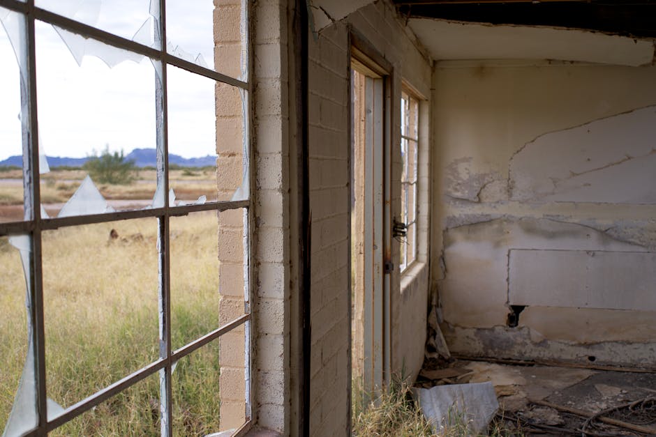 Interior of an abandoned house with broken windows and peeling walls, surrounded by rural landscape.